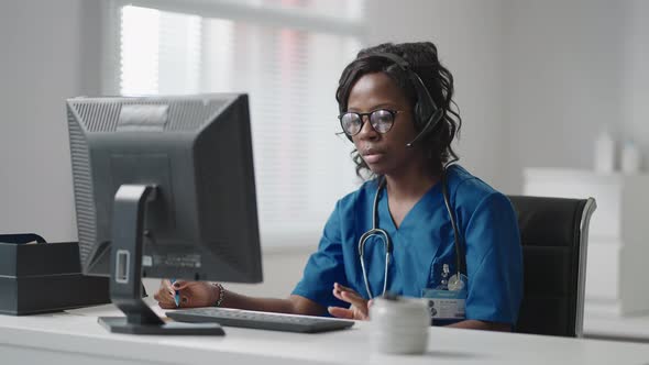 African American Black Female General Practitioner in White Coat Sitting at Desk in Doctor's Office