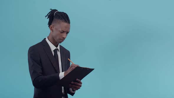 Black Office Man in a Suit and with Dreadlocks Holds a Paper Tablet and Thinking Takes Notes in