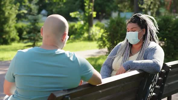 Two Happy Friends in Medical Mask Keep Social Distance Sitting and Talking Outside on Bench in Park