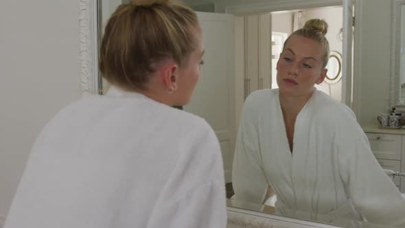 Caucasian woman in bathrobe touching her face while looking in the mirror in the bathroom