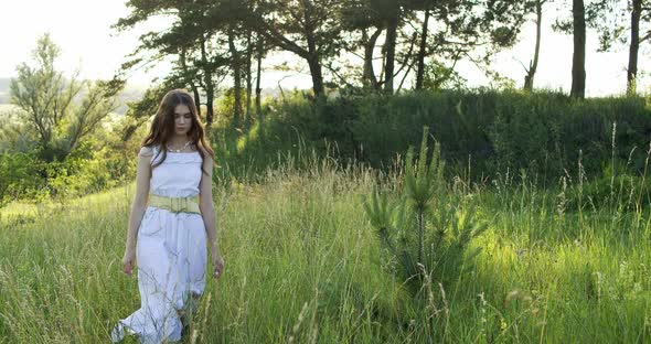 A Cheerful Teenage Girl in Light Dress Walks in a Clearing Between Green Trees in the Summer