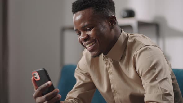 Cheerful Black Man is Talking By Video Call in Smartphone Sitting on Couch at Home Alone Medium