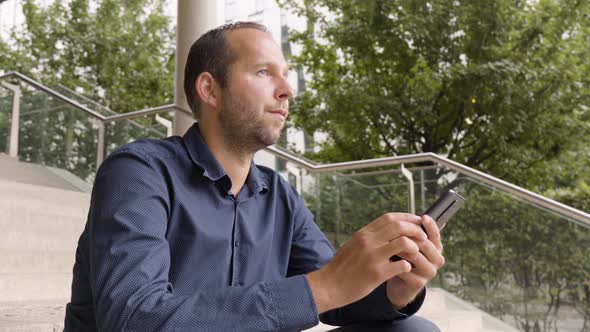 A Caucasian Man Looks Around and Holds a Smartphone As He Sits on a Staircase in an Urban Area