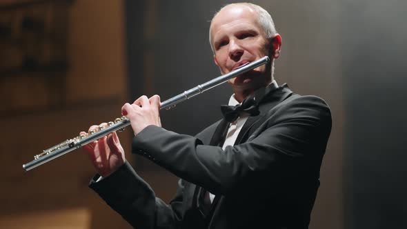 Grey Haired Flutist is Playing Classic Music By Flute Portrait of Musician in Dark Hall of Opera