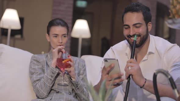 Man And Woman Smoking Shisha, Drinking Cocktails And Using Phone