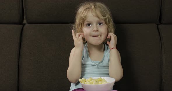 Girl Sitting on Sofa and Eating Corn Puffs. Child Watching Tv, Taste Puffcorns