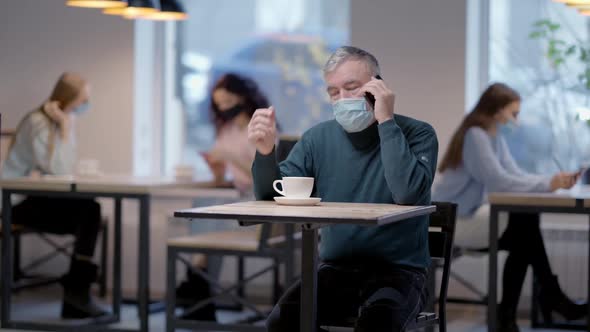 Senior Man in Coronavirus Face Mask Talking on the Phone Sitting in Restaurant with Blurred People