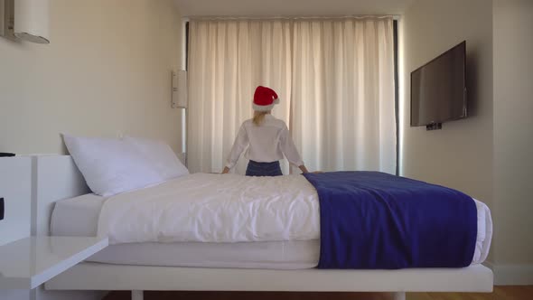 a woman in a Santa Claus hat gets out of bed and opens the curtains.