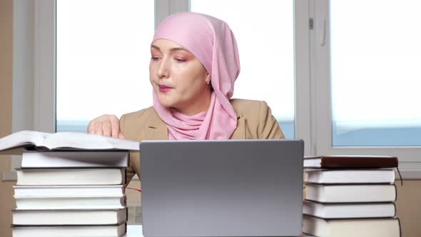 Woman in Hijab Typing on Laptop Among Stacks of Books