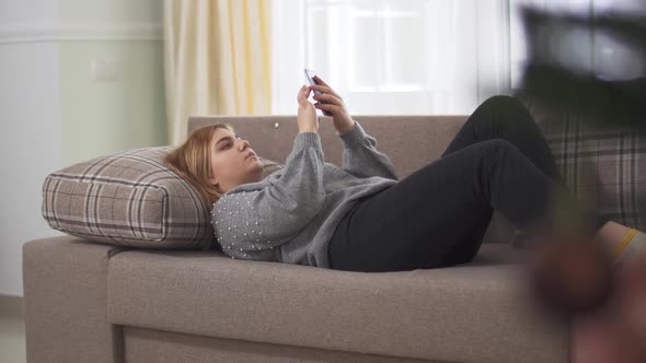 Plus Size Young Woman Texting on Cell Phone Indoors, Lying on the Sofa Near Big Window. Cute Plus