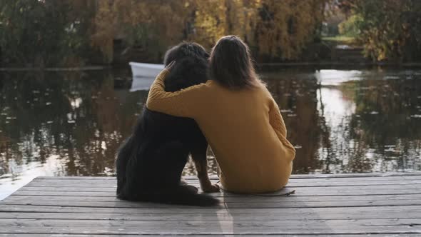 A Young Woman Sits on a Pier Near the River with a Big Dog and Hugs Her. A Boat with People Floating