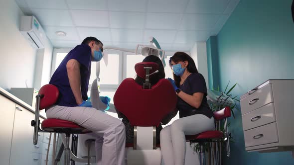 Woman Dentist in Medical Mask with Assistant Examines Patient in Dental Clinic