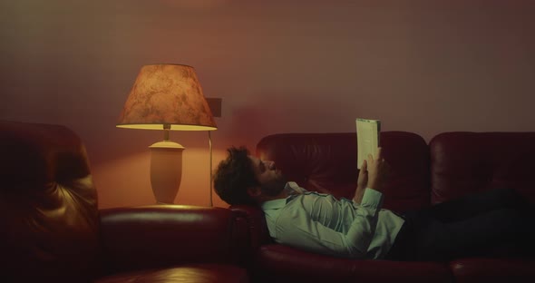 Guy with shirt reads a book lying on the sofa at home