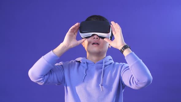 Joyful Japanese Youngster Playing Videogame in Virtual Reality Helmet on Dark Blue Background.