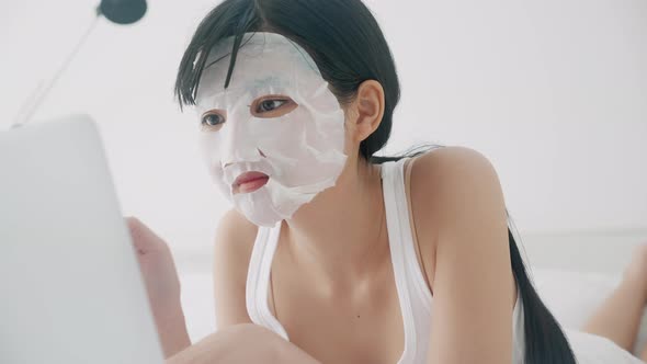 Beautiful young asian woman with sheet facial mask lying working on laptop computer on bed.