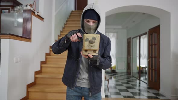 Confident Masked Caucasian Young Burglar Standing in Wealthy House Opening Decorative Box with Gun