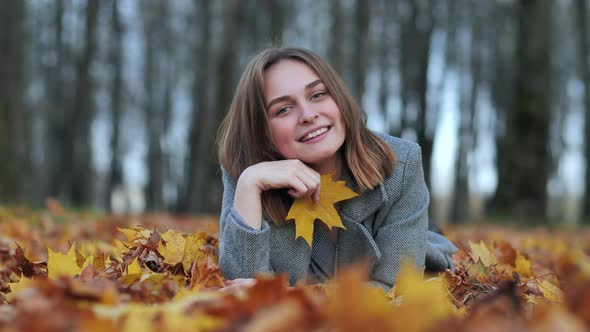 A Young Girl in an Autumn Park Lies on the Leaves and Smiles with a Leaf in Her Hand.
