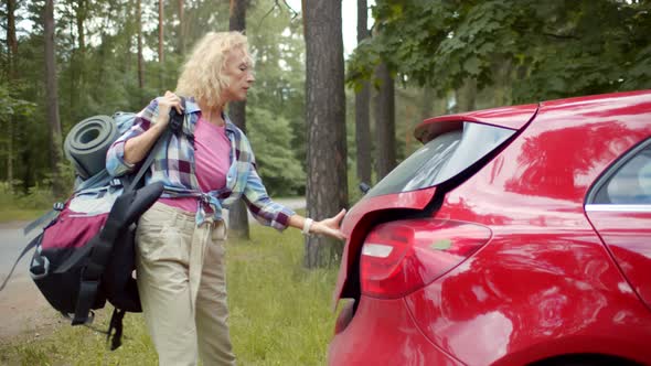 Side View Pf Attractive Mature Woman Loading Hiking Backpack in Red Car in Forest