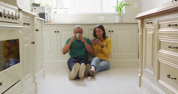 Happy biracial couple sitting on kitchen's floor, drinking coffee and talking