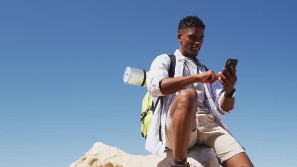 African american man hiking using smartphone sitting on rock by the coast