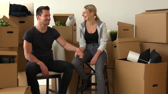 A Happy Moving Couple Sits on Chairs, Holds Hands and Smiles at the Camera in an Empty Apartment