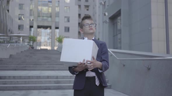 Cute Little Boy in a Business Suit and Glasses Holding a Box with Stationery on the Stairs Outdoors