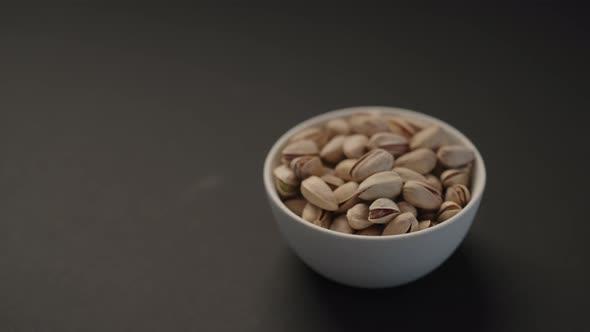 Handheld Slow Motion Salted Large Pistachios in White Bowl on Black Paper Background