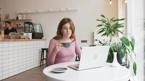 Beautiful Young Redhead Woman Working with Laptop in Cafe and Drinking Coffee