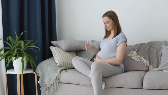 Pregnant Woman in Home Clothes Sitting on the Couch Holding a Glass of Water in Her Hands