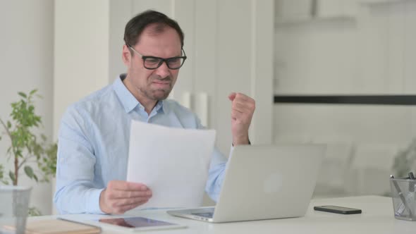 Upset Middle Aged Man Reading Documents While Using Laptop