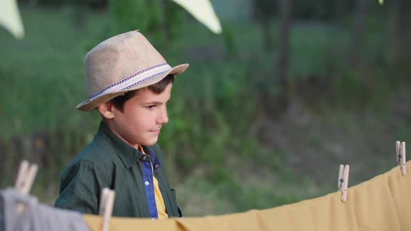 Adorable Boy in a Hat Makes a Wigwam and Clings Clothespins While Relaxing Outdoors on a Summer