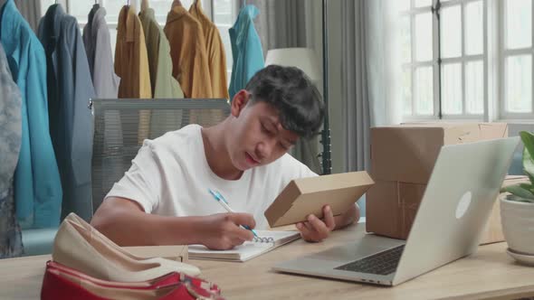 Asian Man Online Seller Looking At The Package And Writing In The Notebook While Selling Home