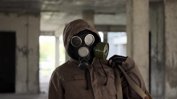 Man Wearing Gas Mask and Postapocalyptic Old Scruffy Clothes in Abandoned Building Puts Backpack on