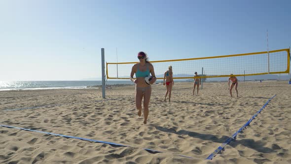 Women players play beach volleyball and a player serves the ball.