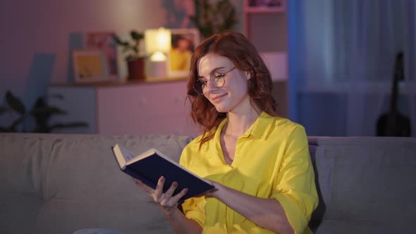 Woman with Glasses for Sight Enjoys Reading Book While Sitting on Couch in House in Evening Stay at