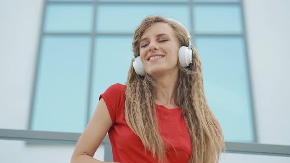 A Young Girl in Headphones is Listening to Music and Amusingly Dancing on a City Background