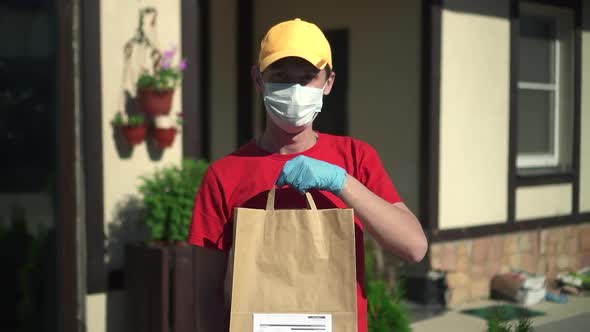 Young Man Courier Poses with Paper Bag in His Hands on Background of House Outdoors Spbd