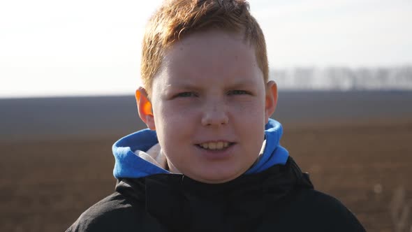 Serious Kid with Freckles Looks Into Camera Against the Blurred Background of Plowed Field