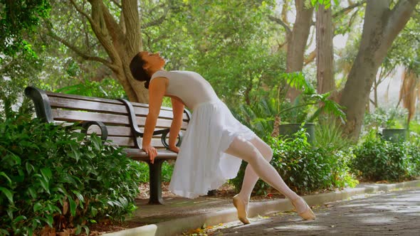 Beautiful young ballerina doing stretching exercise on bench in the park 4k