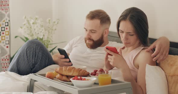 Young Couple Using Their Smartphones While Lying in Bed in Morning, Man and Woman Looking at Phone