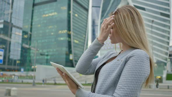 A Young Blond Girl Uses a Tablet on a Background of Skyscrapers Downtown