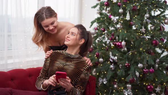 Two Women are Sitting on a Sofa Near the Christmas Tree Looking at the Screen of a Smartphone