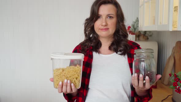 a Beautiful Pregnant Woman in a Red Shirt in the Kitchen Holds a Jar of Pasta and a Jar of Red Beans