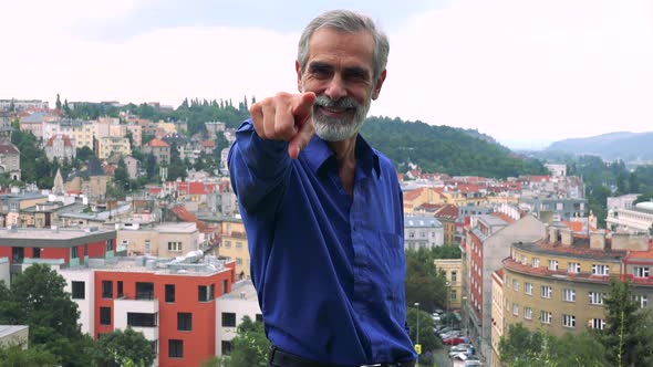 Old Senior Man Points To Camera with Finger - City (Buildings) in Background