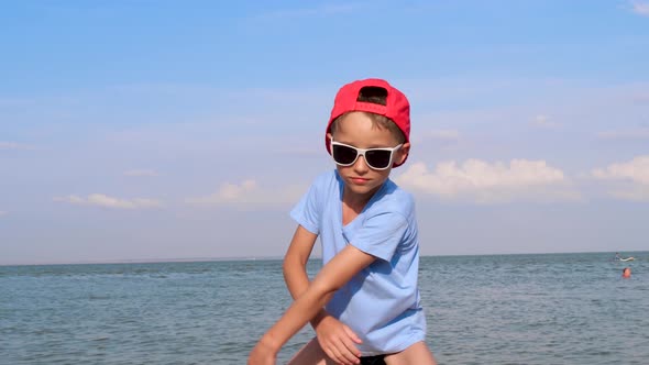 A Cheerful Child in Sunglasses and a Baseball Cap is Dancing on the Beach