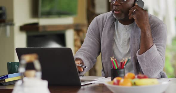 Thoughtful african american senior man wearing bathrobe using laptop and talking on smartphone