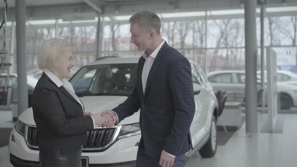 Portrait of Caucasian Car Dealer Welcoming Senior Female Client in Dealership. Wealthy Blond Woman