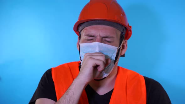 Male Construction Worker in Overalls and Medical Mask Coughing on Blue Background