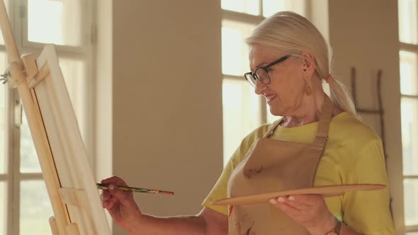 Happy Senior Woman Artist Paints a Picture on Canvas While Standing in a Sunny Workshop