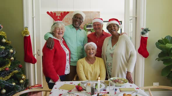 Portrait of diverse senior friends in christmas hats embracing and smiling at christmas dinner table